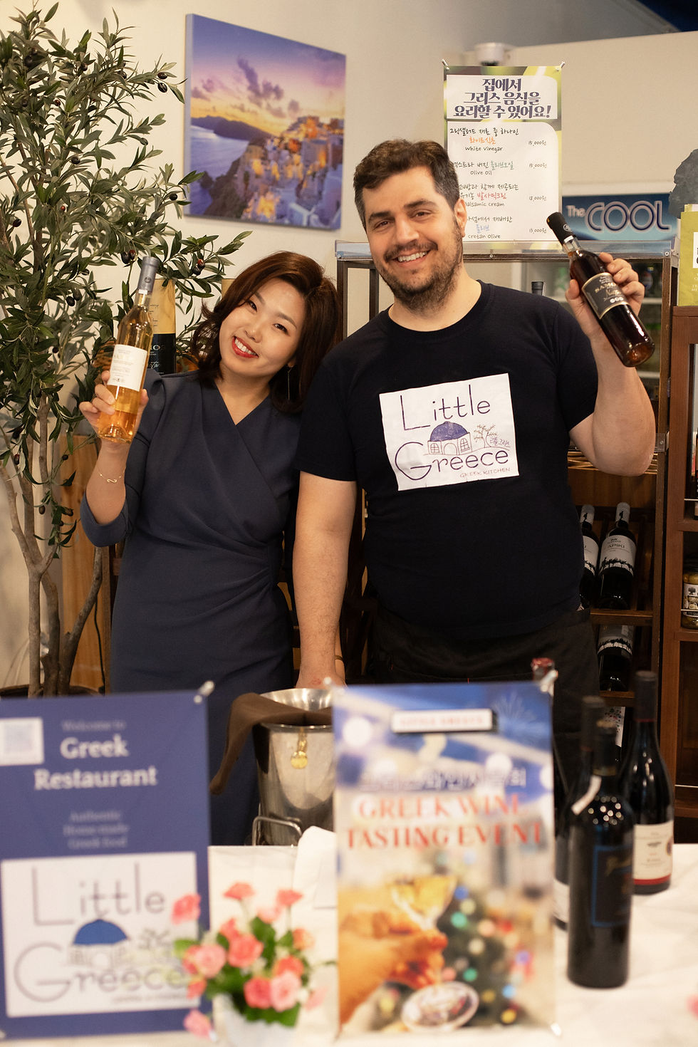 Two people smile while holding wine bottles at a Greek wine tasting event. A sign reads "Little Greece." An olive tree and flowers decorate the scene.