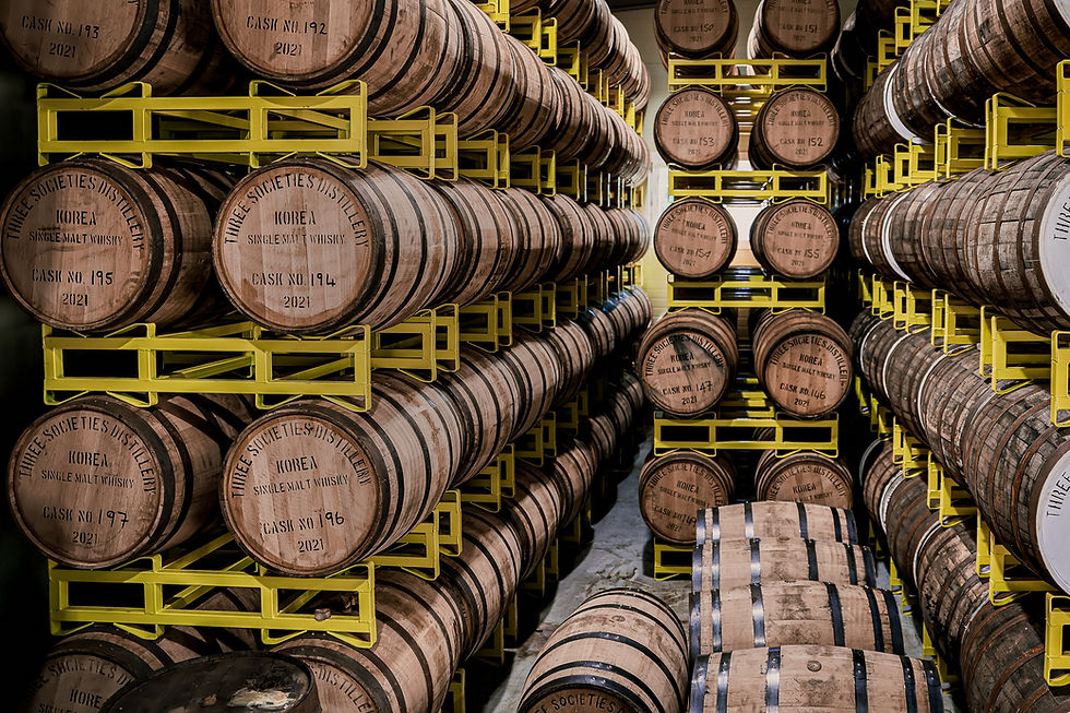 Wooden whiskey barrels stacked in rows on yellow racks in a distillery. Some barrels labeled "Three Societies Distillery, Korea, 2021."