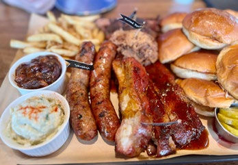 Assorted BBQ platter with ribs, sausages, pulled pork, buns, fries, and sides. Labels on meat. Warm, inviting presentation on a wooden table.
