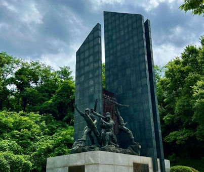Monument with bronze figures in dynamic poses on a stone base. Tall, dark panel backdrop. Cloudy sky, lush green trees in background.