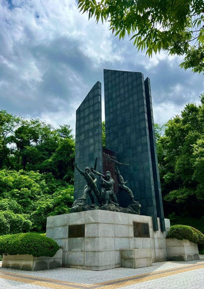 Monument with bronze figures in dynamic poses on a stone base. Tall, dark panel backdrop. Cloudy sky, lush green trees in background.