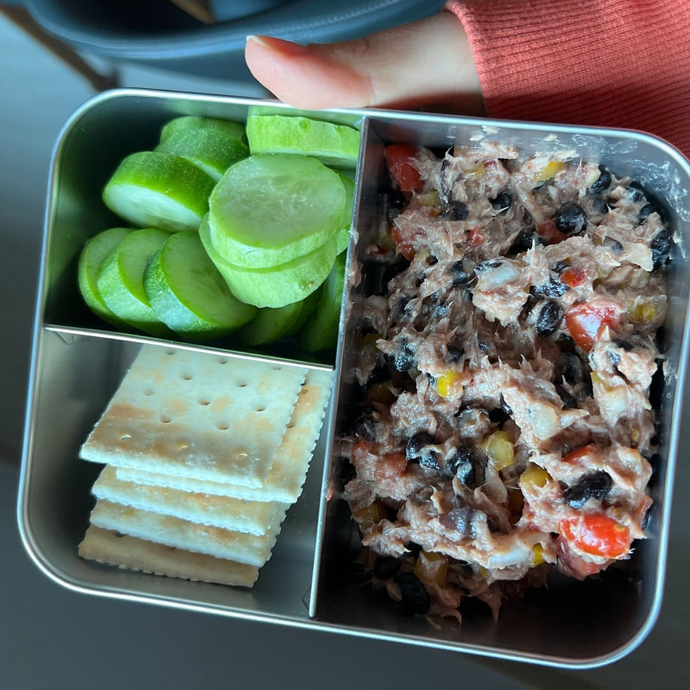 Bento box with cucumber slices, crackers, and a colorful tuna salad. A hand in an orange sleeve holds the box. Bright, casual setting.