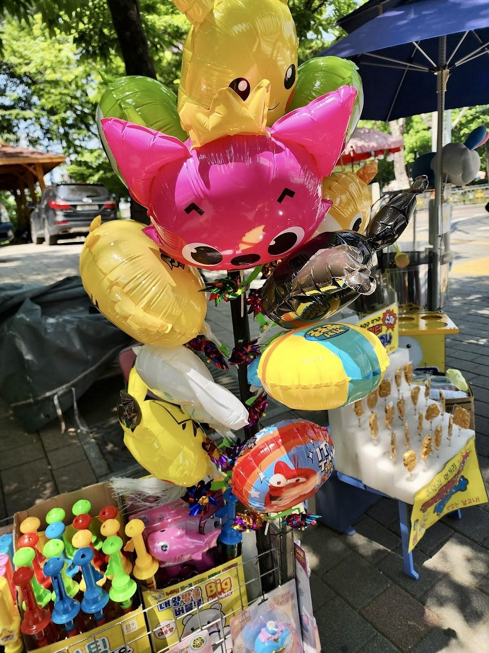 Colorful animal-shaped balloons tied to a stand with toys and snacks nearby. It's a sunny day on a sidewalk with trees and cars. Text visible.