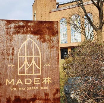 Rusty sign reading "MADE 森 YOU MAY DREAM HERE" in front of a brick building with arched windows and bare tree branches.