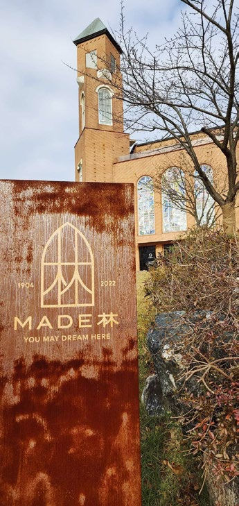 Rusty sign reading "MADE 森 YOU MAY DREAM HERE" in front of a brick building with arched windows and bare tree branches.