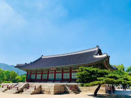 Traditional Korean building with a curved roof in a sunny park setting. Clear blue sky, green trees, and people walking nearby