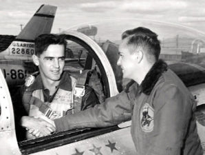 Two men in military uniforms, one in a jet cockpit shaking hands with another standing outside. A plane tail number and patches visible.