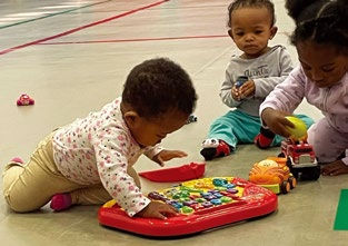 Three toddlers play on a gym floor with colorful toys, including a musical keyboard and a toy truck. They appear focused and curious.