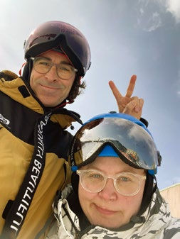Two people in ski gear smile for a selfie. One flashes a peace sign. Blue sky in the background. Mood is playful and cheerful.