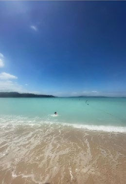Person swimming in clear turquoise sea under a bright blue sky. Sandy beach in the foreground. Calm, serene atmosphere.