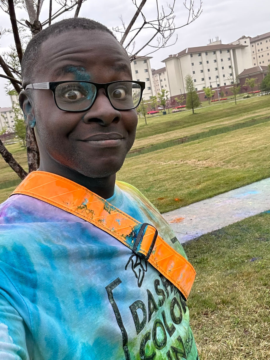 Person with colorful paint on face and shirt smiles in a park with buildings in the background. Shirt text reads "Dash Color Run."