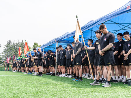 A group of people in black shorts and shirts labeled "ARMY" stand solemnly on a green field under blue canopies, holding flags.