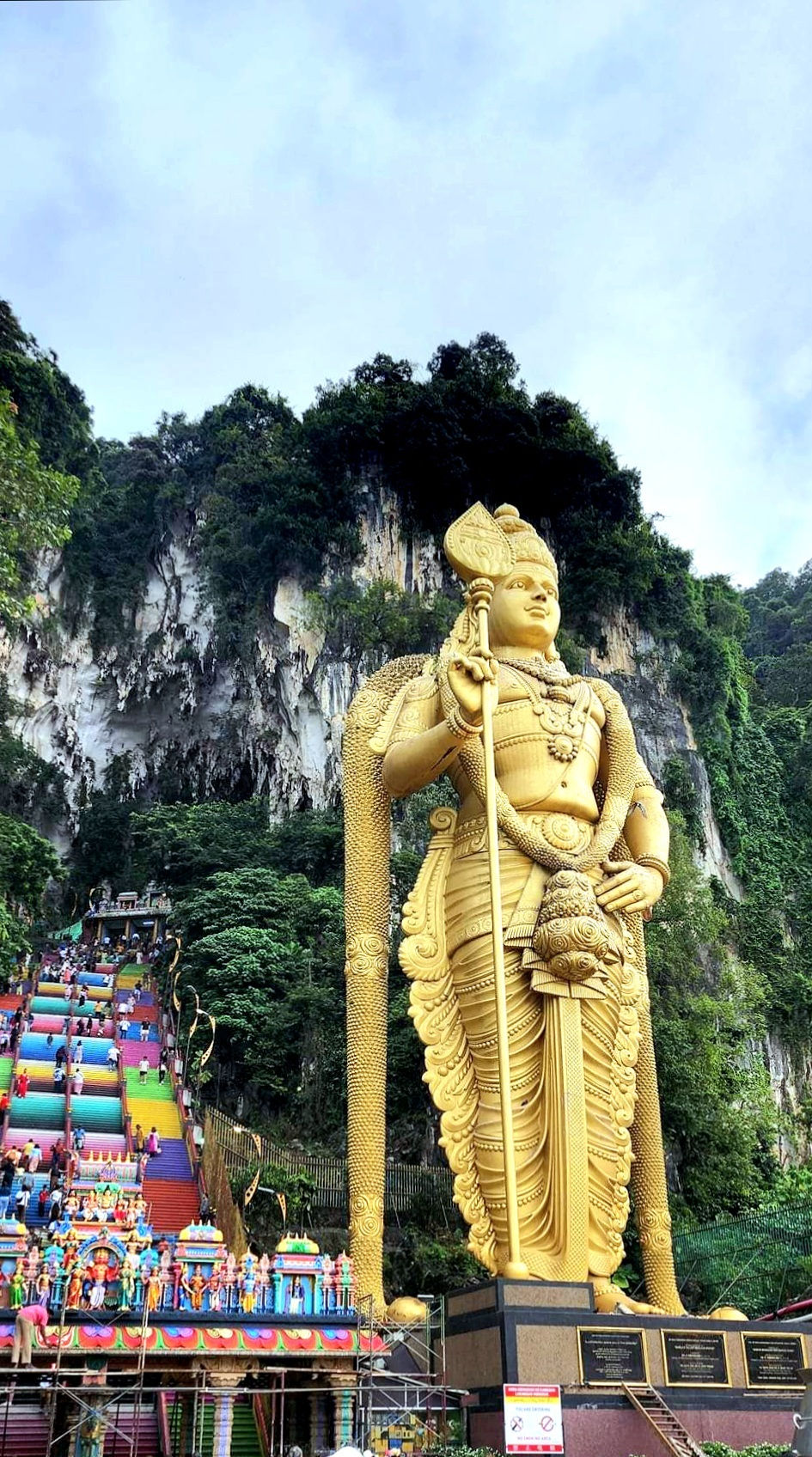 Giant golden statue of a deity beside colorful stairs with people climbing. Lush green cliffs and sky in the background. Peaceful mood.