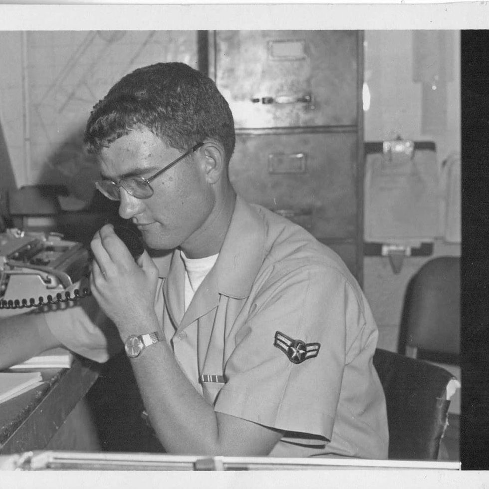 Young man in uniform speaking into a microphone at a desk, with a typewriter and file cabinet in the background. Serious expression. Black and white.