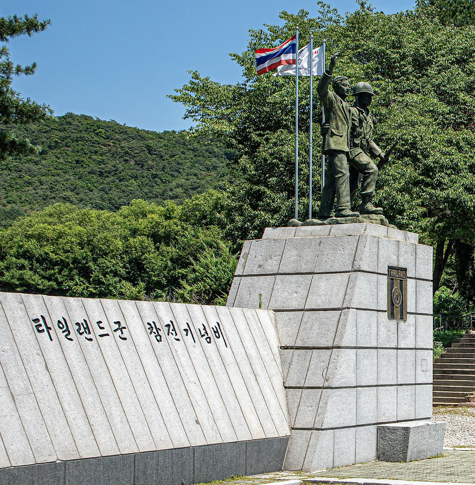 Statue of two soldiers on a stone pedestal with Thai and Korean flags, set against a green, tree-filled background. Korean text visible.