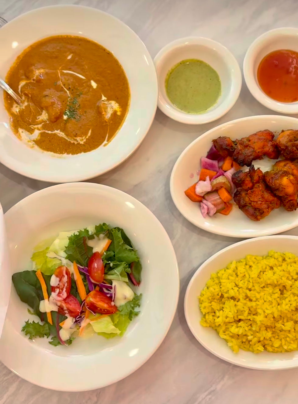 Plates of curry, yellow rice, salad, grilled chicken, and sauces on a marble table. Vibrant colors and appetizing presentation.