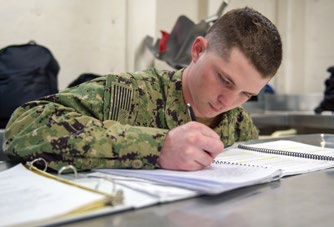A person in military uniform writes in a notebook on a table, focused and serious. Background shows bags and a neutral wall.