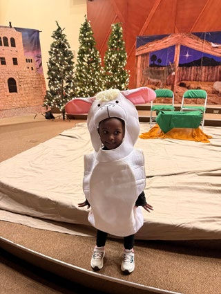 Child in a sheep costume stands on a stage with Christmas trees and nativity scene backdrop. Warm lighting and festive atmosphere.