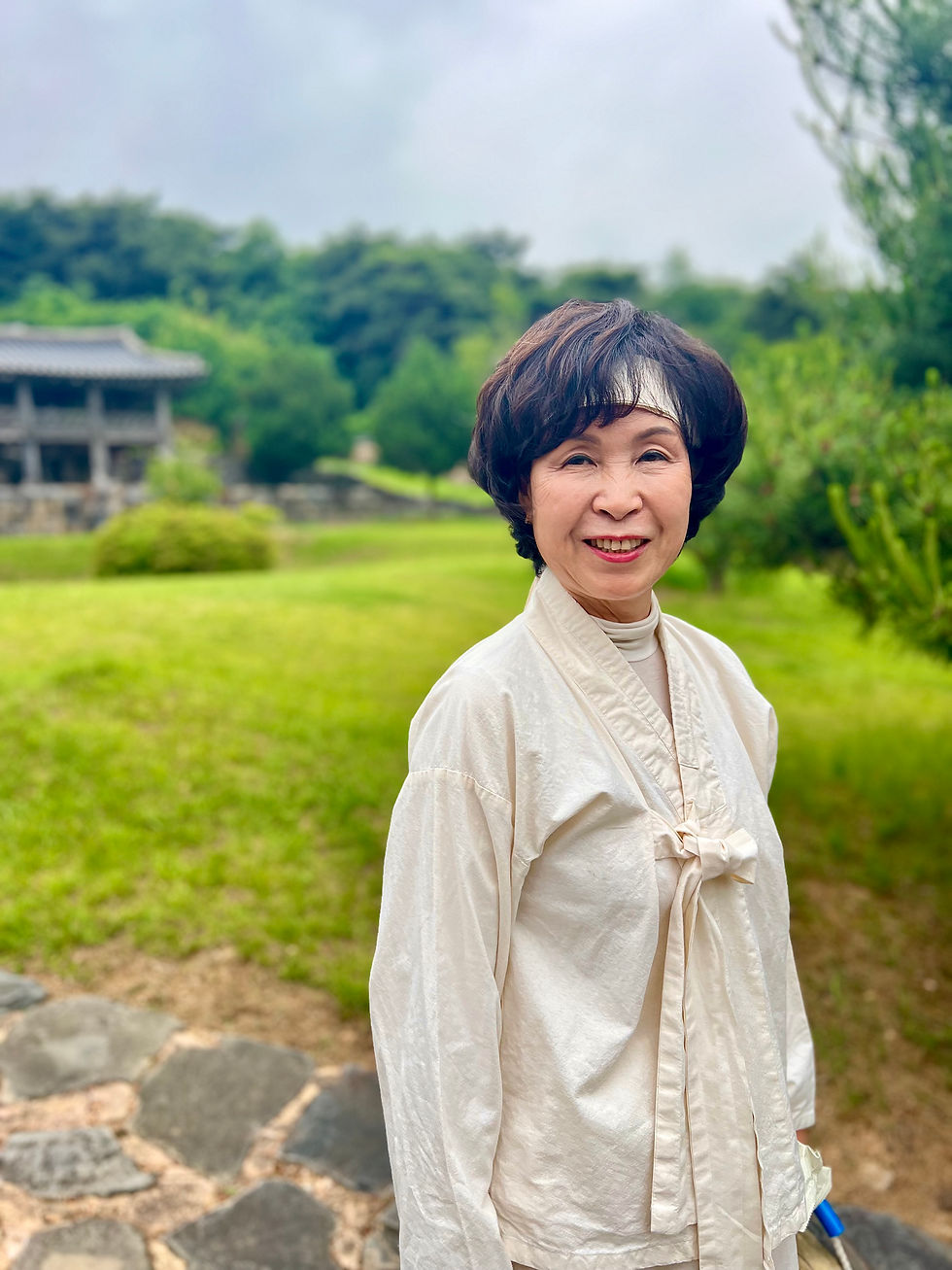 Smiling woman in a white traditional outfit stands on a stone path in a lush garden. Overcast sky, old building in the background.