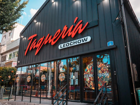 Black building with red "Taqueria" sign, windows displaying colorful logos. "LEOCHOW" and "open" visible. Sunny day, urban setting.
