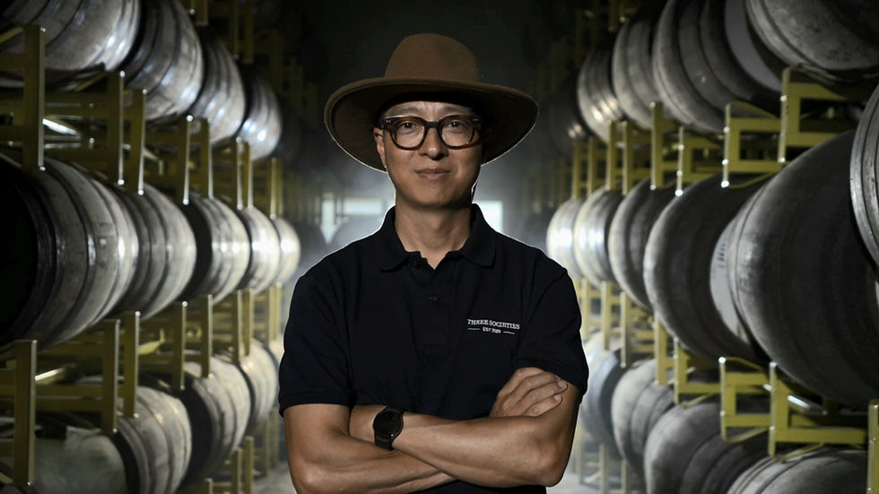Man in a hat stands confidently with arms crossed in a dimly lit warehouse, surrounded by rows of stacked barrels. Wearing a "Three Societies" shirt.