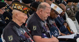 Veterans in uniformed hats sit solemnly with papers at an event. Military personnel in the background. Flags and medals visible.