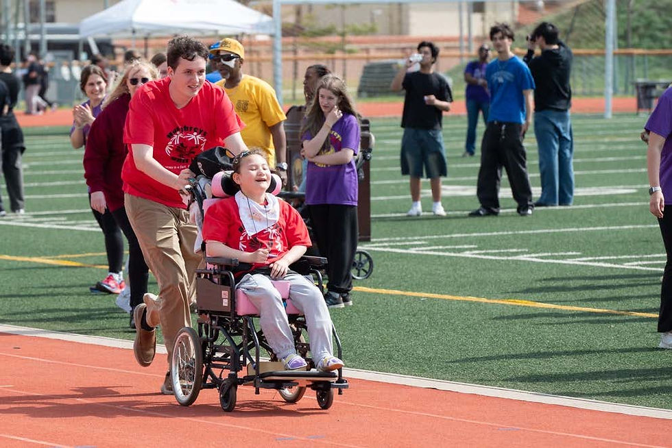 Man in red shirt pushes a smiling child in a wheelchair on a track. Others in colorful shirts watch and cheer. Bright, sunny day.