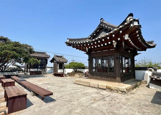 Traditional wooden pavilion in a courtyard under a clear blue sky. Benches and trees in the background suggest a serene atmosphere.