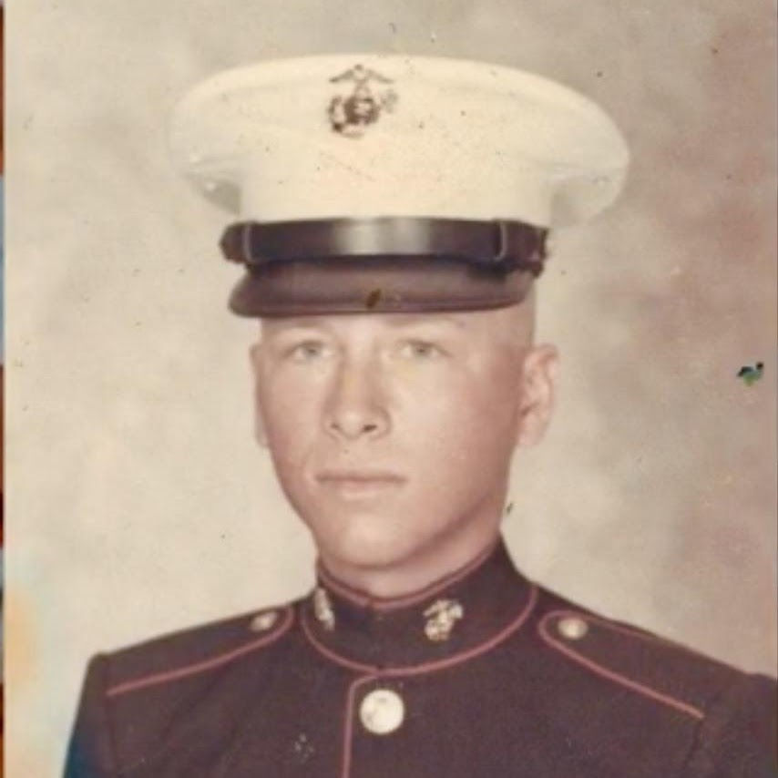 Young man in a Marine dress uniform, white cap with emblem, neutral expression. Background is plain and beige. No visible text.