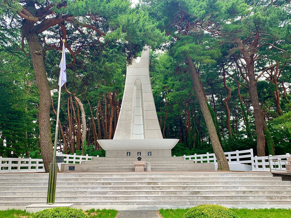 A white monument with a sleek design stands among tall pine trees. A flag on a pole is nearby. The atmosphere is serene and solemn.