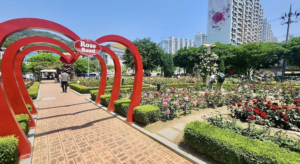 Red heart arches labeled "Rose Road" adorn a flower garden. People stroll along brick paths, surrounded by colorful roses and green hedges.