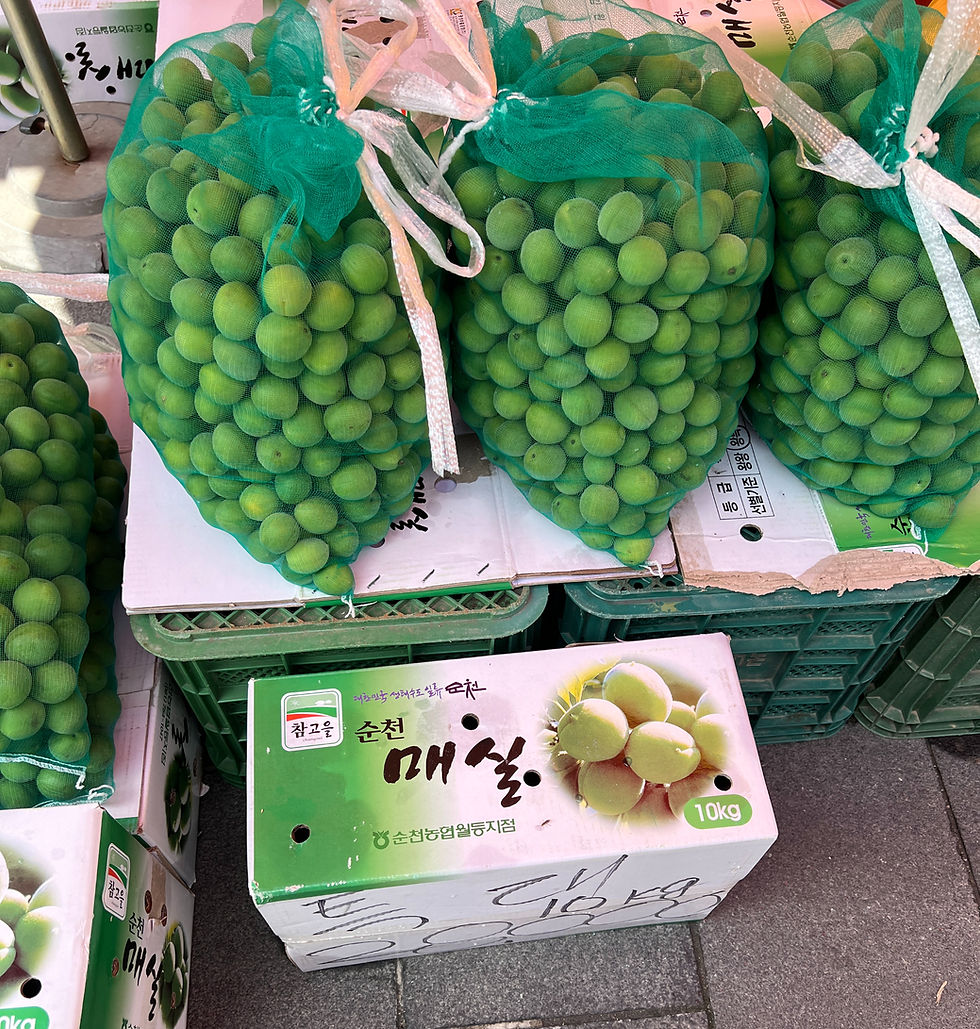 Green plums in net bags on boxes, text includes "10kg." The setting is a street market with a tiled floor. Bright and fresh mood.