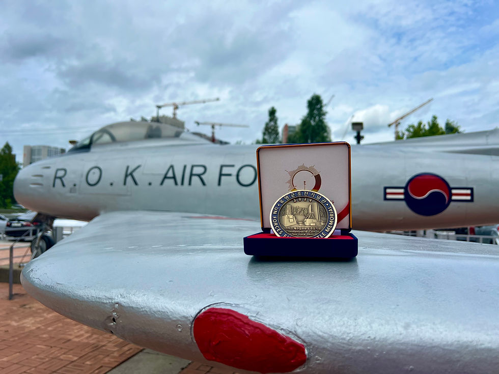 A commemorative coin in a case on an aircraft's wing, labeled "R.O.K. AIR FORCE," under a cloudy sky with cranes in the background.