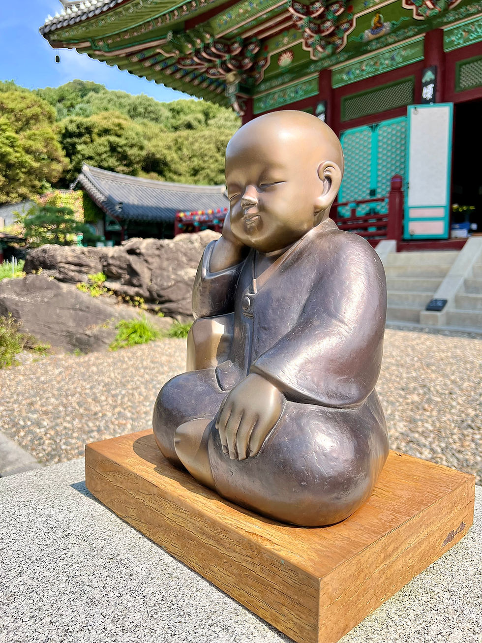 Bronze statue of a meditating monk child in a serene pose on a wooden base. Traditional temple with ornate roof in background; lush greenery.