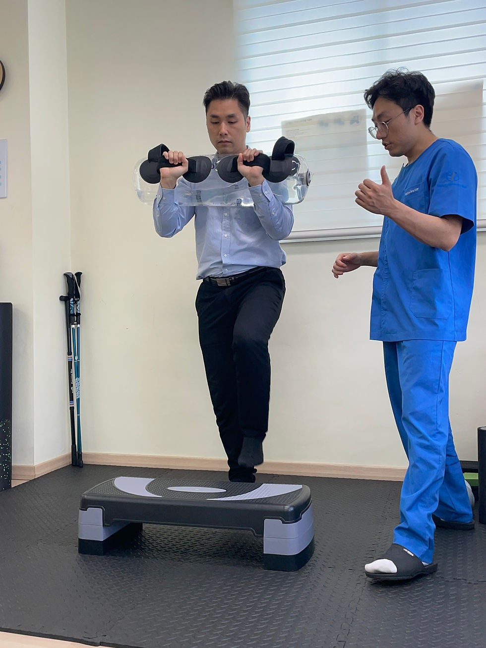 Man in blue shirt balances on step with a water weight while another man in blue scrubs observes in a neutral-colored room.