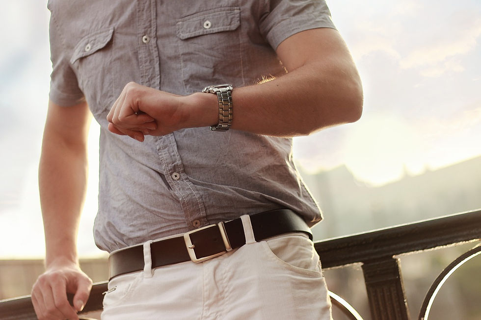 Person in a gray shirt and white pants checks silver watch. Sunny background with railing, casual and slightly hurried mood.