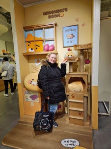 Woman in a black coat poses smiling in front of a cartoon-themed display titled "CHOONSIC'S ROOM" with cat artwork in a store setting.