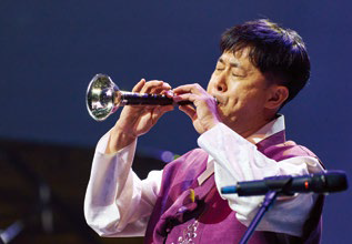 Man in a purple vest passionately plays a trumpet-like instrument on stage, eyes closed. Blue background with a microphone nearby.