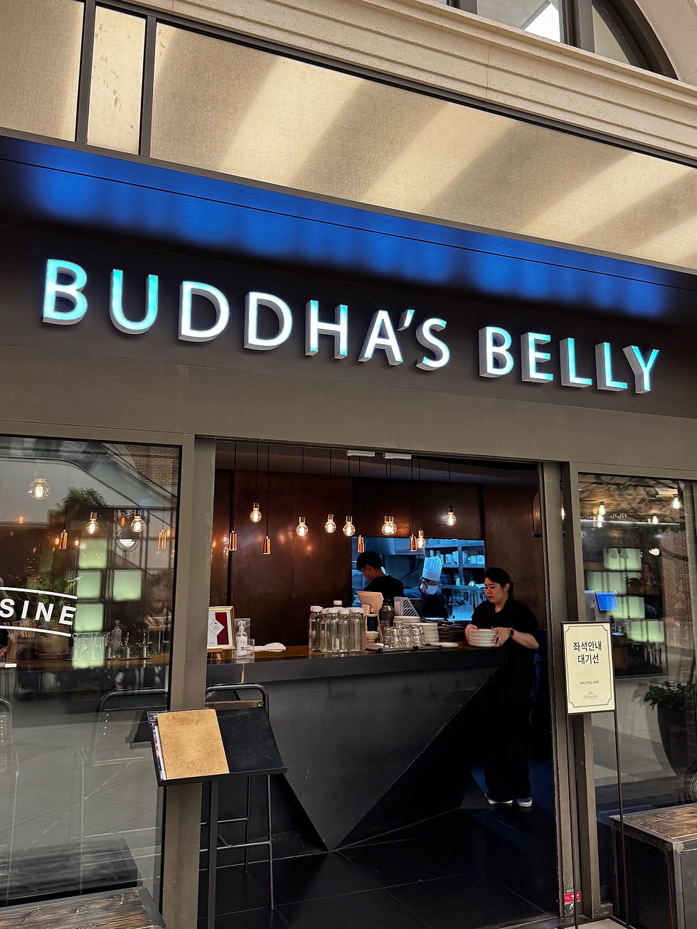 Restaurant front labeled "Buddha's Belly." Inside, staff prepare food under warm pendant lights. Modern, inviting atmosphere.