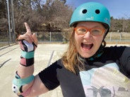 Woman in teal helmet and red glasses excitedly points upwards at skate park. Wearing black shirt with graphic; colorful wrist guards. Blue sky.
