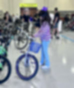 Girl in striped shirt and purple bow stands with a bike indoors. Bicycles and people in the blurred background. Calm atmosphere.