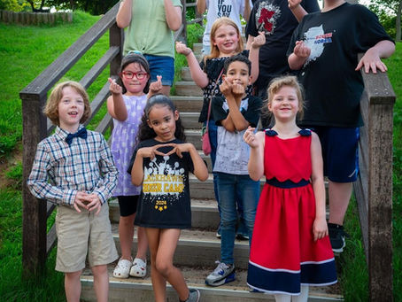 A group of smiling kids and teens on outdoor steps, making heart gestures. They're dressed casually with vibrant colors in a green park setting.