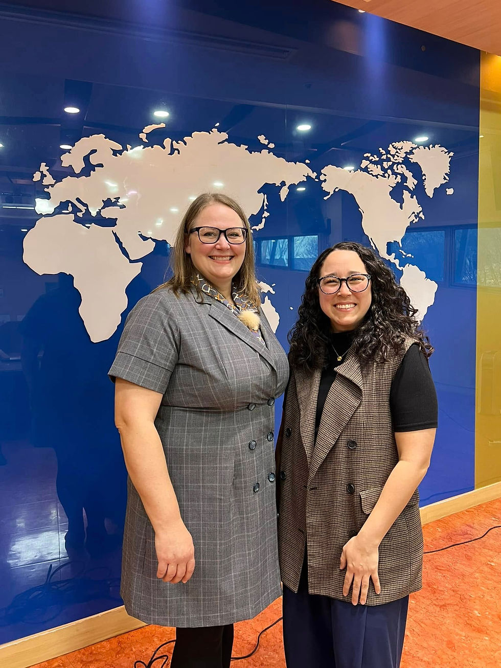 Two women smiling in front of a blue wall with a world map. Both wear glasses and blazers, exuding a professional and cheerful mood.