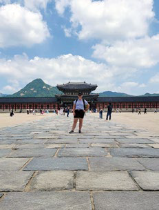 Person standing on stone path at Gyeongbokgung Palace, Seoul. Bright day, blue sky, clouds, and mountain backdrop. Historical ambiance.