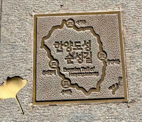 Bronze plaque on a stone path reads "Excursion Trail of Hanyangdoseong" with Korean text; a yellow ginkgo leaf lies nearby.