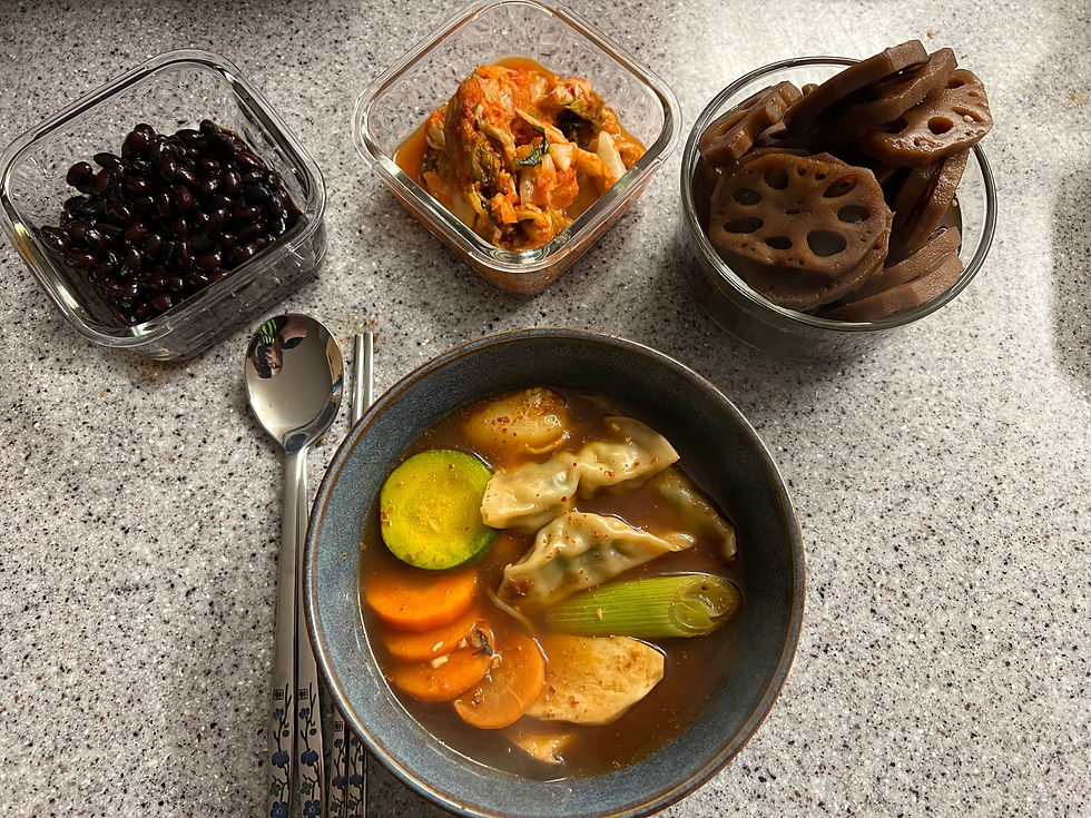 Bowl of soup with dumplings, zucchini, and carrots on a speckled surface. Dishes of black beans, kimchi, and lotus root slices nearby.