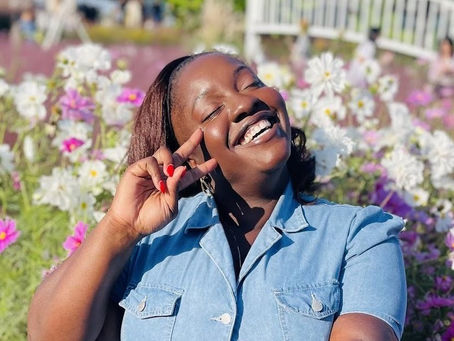 Smiling woman in a blue dress poses with peace sign in a vibrant flower garden, sunny day. Bright flowers and greenery in the background. Military Spouse Work Life Balance