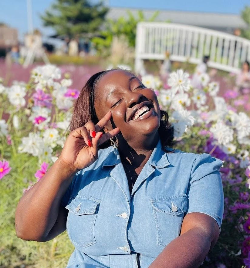 Smiling woman in a blue dress poses with peace sign in a vibrant flower garden, sunny day. Bright flowers and greenery in the background. Military Spouse Work Life Balance