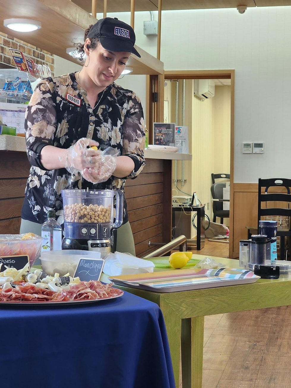 A woman in a floral blouse and cap prepares food with a blender at a counter, surrounded by snacks and ingredients. Cozy kitchen setting.