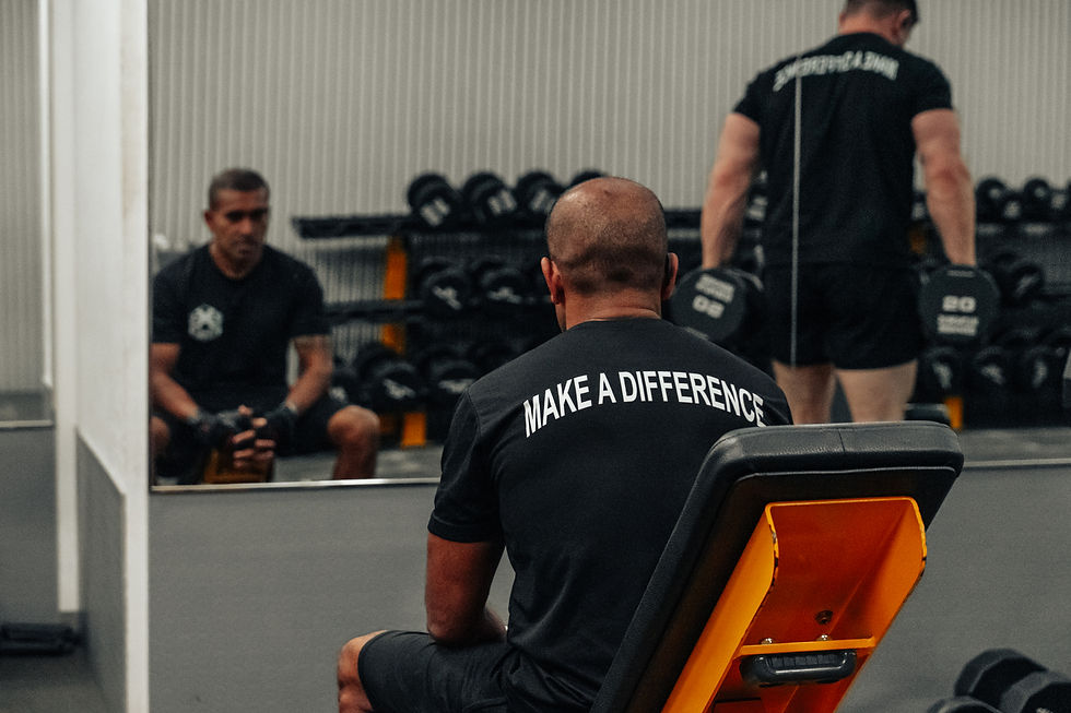 Two men in a gym, one seated with "MAKE A DIFFERENCE" on his shirt, another lifting dumbbells. Mirror reflection shows focused mood.
